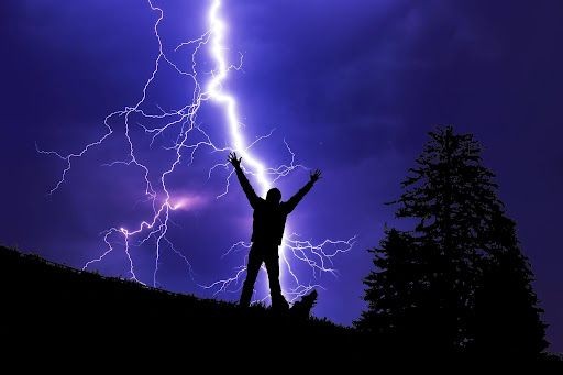 Silhouette of person with raised arms against dramatic lightning in purple sky, next to trees.